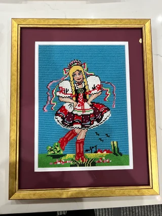 Needlework figure in a gold frame with burgundy and white matting.